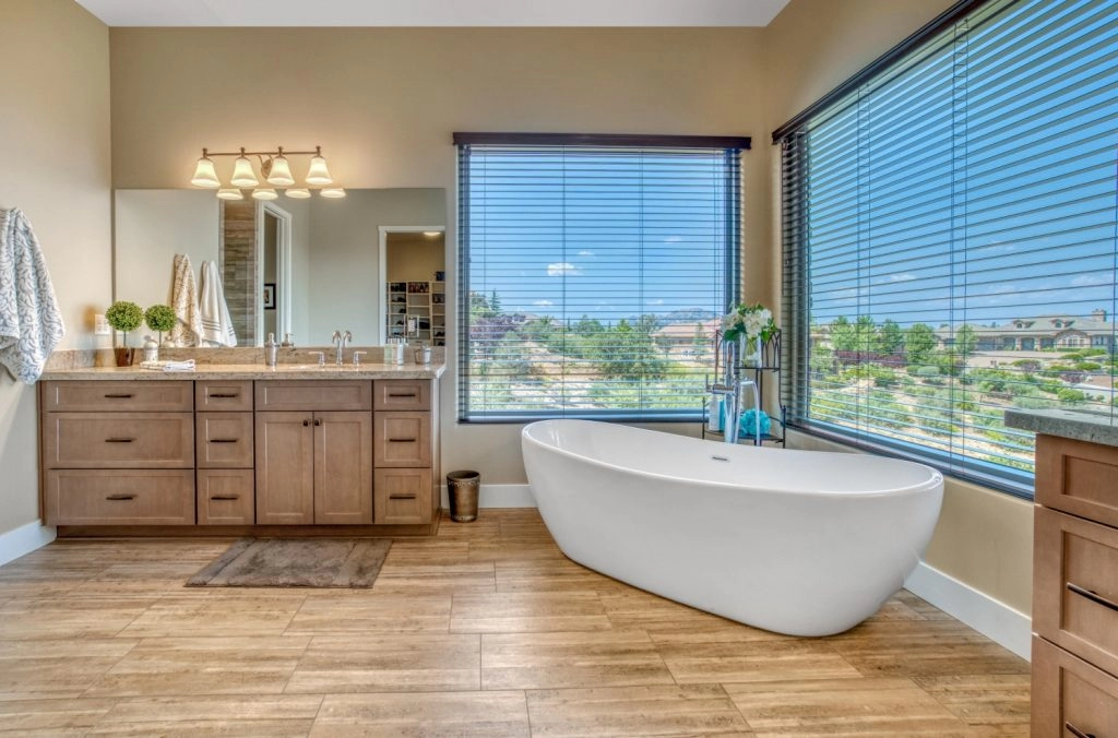 Bright and spacious luxury bathroom featuring a freestanding tub, wooden cabinetry, and large windows with scenic views.