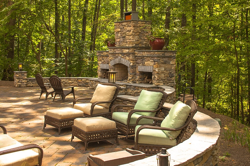 Beautiful stone patio with comfortable chairs and a fireplace surrounded by lush green trees.