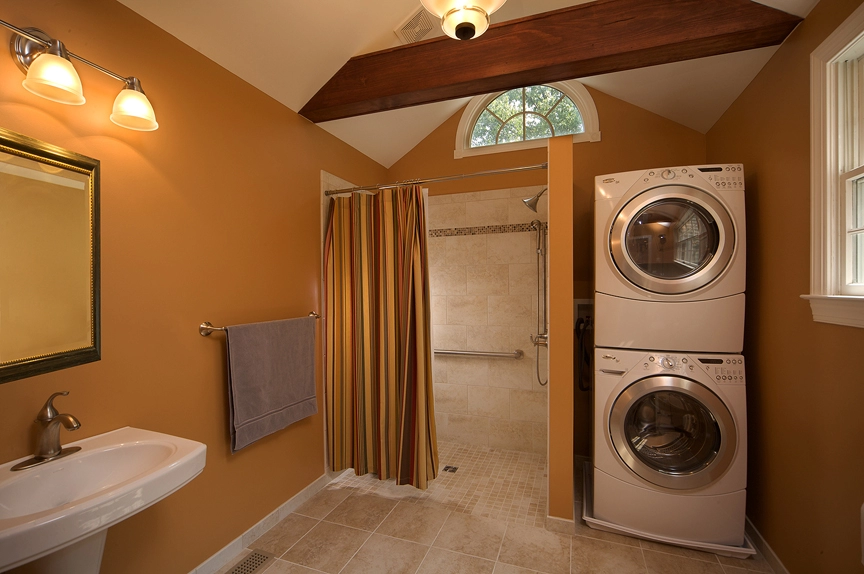 Modern bathroom with a stacked washer and dryer, beige tile flooring, and warm orange walls, featuring a shower area.