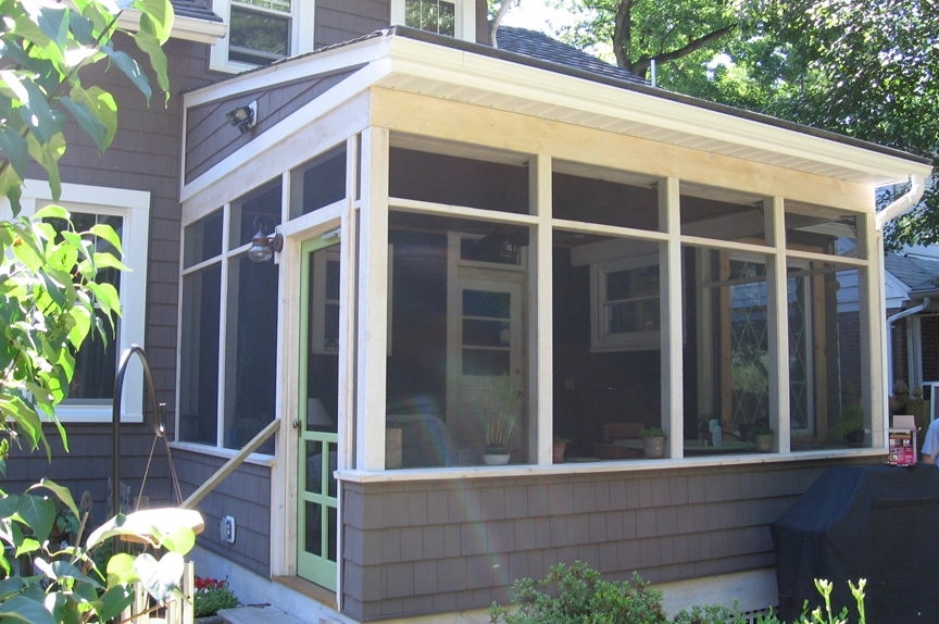 A screened-in porch featuring large windows, wooden siding, and a green door, perfect for outdoor relaxation.