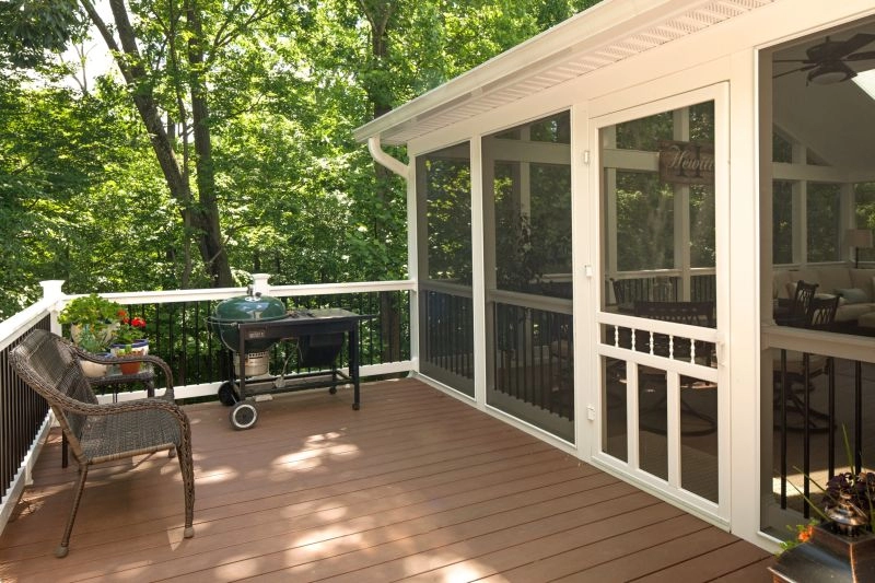 Spacious screened porch featuring a wooden deck, outdoor grill, and cozy seating surrounded by lush greenery.