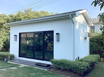 Modern outdoor gym shed featuring white paneling and large black glass doors, surrounded by green landscaping.